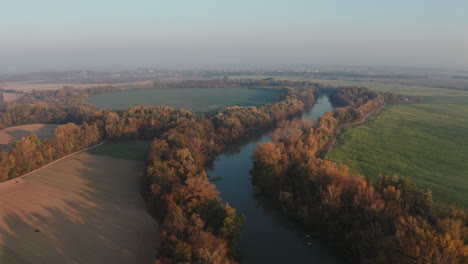 Wide-angle-Aerial-shot-of-small-Danube-river-with-two-white-birds-flying-over-and-Colorful-Autumn-forest,-fields