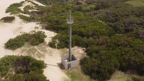 Aerial-oribit-shot-of-telecommunication-tower-on-sandy-beach-and-green-bush-dunes-during-sunny-day---Mar-de-las-Pampas,Argentina