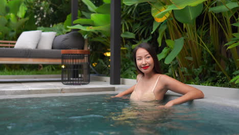 Fröhliche-Asiatische-Frau-Im-Hautfarbenen-Bikini,-Die-In-Einem-Luxuriösen-Swimmingpool-In-Einem-Modernen-Tropischen-Spa-Bereich-Im-Freien-Genießt,-Mittlere-Aufnahme-Mit-Kopierraum