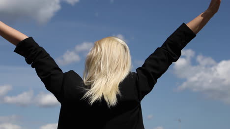 Back-view-of-blond-businesswoman-is-standing-with-raised-arms-on-blue-sky-background
