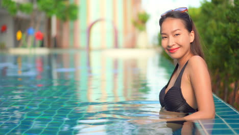 An-attractive-Asian-woman-lounging-in-the-shallow-end-of-a-resort-pool-smiles-for-the-camera