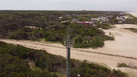 Aerial-orbit-shot-of-network-antenna-at-beach-and-forest-of-Mar-de-las-Pampas-sending-signal-waves-to-city,Argentina