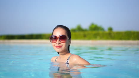 Sexy-Asian-woman-with-big-sunglasses-standing-in-swimming-pool,-smiling-to-the-camera