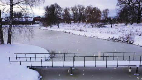 Small-city-park-with-bridge-over-frozen-lake-water-in-winter-season-during-snowfall,-aerial-view