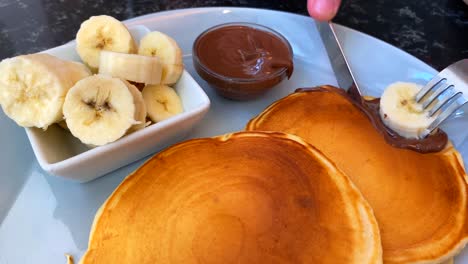 A-person-cutting-golden-pancakes-with-bananas-and-chocolate-sauce-on-the-side,-tasty-breakfast,-4K-shot