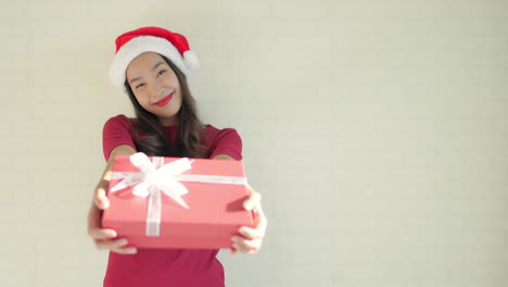 medium-wide-of-isolated-young-adult-Asian-woman-against-off-white-studio-background-with-red-elegant-casual-clothing-and-Christmas-hat-smiling,-offer-wrapped-present-gift-box-Meme-shot-with-copy-space