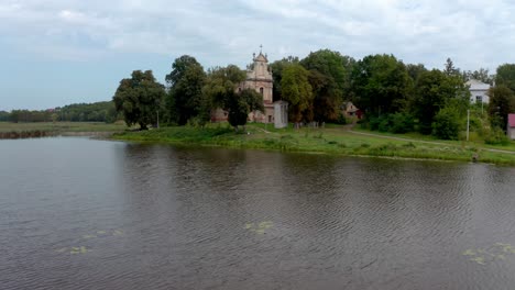 Hodovytsya,-Ukraine---All-Saints-Catholic-Church-Surrounded-By-Green-Trees-Built-Near-The-Placid-Lake-Under-The-Cloudy-Sky---Wide-Shot