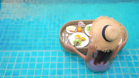 Young-woman-sits-on-edge-of-pool-with-floating-tray-of-food