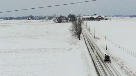 Aerial-tracking-shot-of-Amish-horse-and-buggy-on-rural-country-road-during-winter-snowstorm