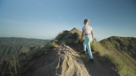 Blond-woman-hiking-on-ridge-on-Mount-Batur-during-morning-trek,-reaching-top-hands-up