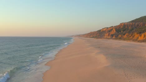 Flying-through-an-empty-beach,-Arriba-Fossil-da-Praia-da-Gale-Fontainhas-seaside-cliff-rock-formation-during-sunset-in-the-coast-shore