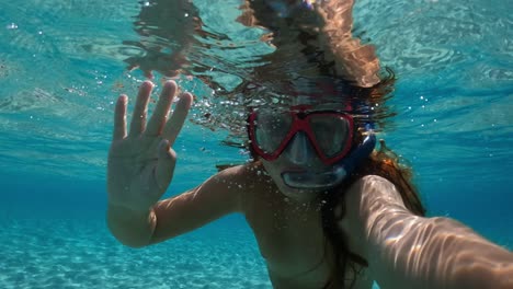 Underwater-selfie-of-little-red-haired-girl-with-diving-mask-and-snorkel-greeting-waving-hand-and-swimming-in-clear-sea-water