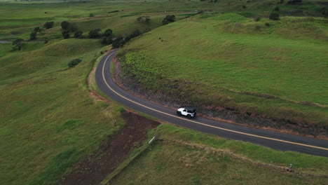 Toma-Aérea-De-Un-Jeep-Conduciendo-Por-Una-Carretera-Con-Curvas-En-Hawai-Con-Un-Exuberante-Paisaje-Verde