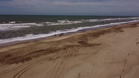 Aerial-shot-of-four-people-riding-horses-along-sandy-beach-with-waves-of-Atlantic-Ocean-during-summer-day