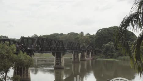 An-establishing-shot-of-the-historic-landmark-the-Bridge-over-the-River-Kwai,-the-water-of-the-Khwae-Noi-river-flowing-calmly-below-the-bridge-in-Kanchanaburi,-Thailand
