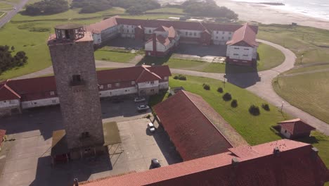 Aerial-orbit-shot-of-old-farming-hotel-with-tower-and-historic-buildings-beside-beautiful-sandy-beach-and-ocean-during-summer---Chapadmalal-in-Argentina