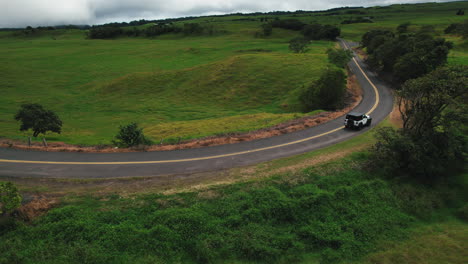 Drone-shot-of-a-white-car-driving-on-scenic-road-with-lush-green-hills-and-trees