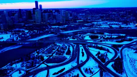 Winter-aerial-drone-fly-out-from-downtown-Edmonton-North-East-Side-to-the-South-East-Side-crossing-the-North-Saskatchewan-River-reflective-shadowed-bridges-spiral-multi-layered-overpasses-into-bridges