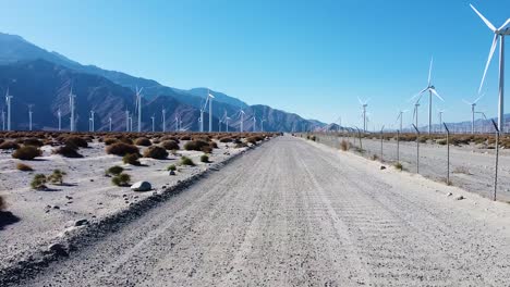 Gravel-roadway-of-massive-wind-turbine-farm-in-California-desert,-aerial-drone-fly-forward-view