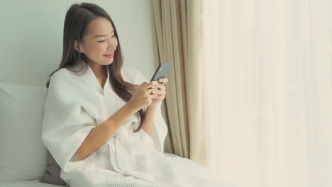 An-attractive-Asian-woman-dressed-in-a-resort-robe-and-sitting-up-on-her-resort-bed-types-a-message-into-her-smartphone
