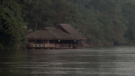 A-distant-shot-of-a-Thai-house-boat-on-the-shore-of-the-fast-flowing-Khwae-Noi-river-in-Kanchanaburi,-Thailand