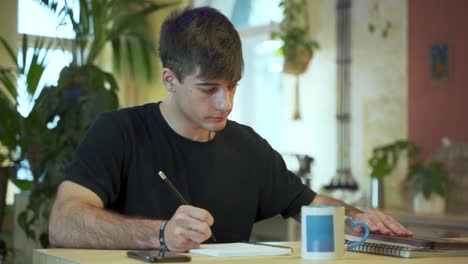 Young-european-Male-Student-is-doing-his-homework-at-the-coffeehouse-with-natural-background