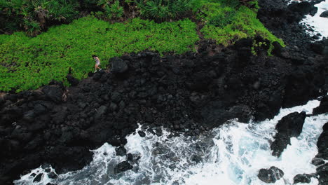 Drone-shot-of-adventurous-young-woman-walking-on-sharp-lava-rocks-over-crashing-waves