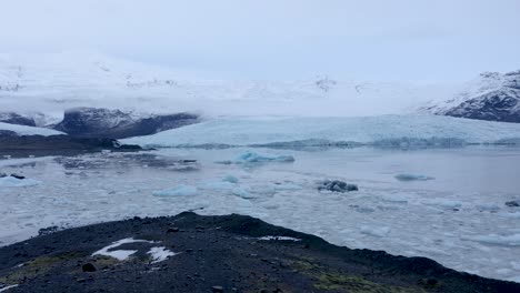 Aerial-forward-flight-showing-icy-Fjallsárlón-glacier-lagoon-in-South-Iceland---Frozen-Vatnajökull-glacier-with-snow,-icebergs-during-cloudy-day