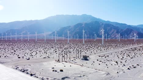 Electric-windmill-farm-in-desert-area-with-majestic-mountain-range-in-background,-aerial-drone-view