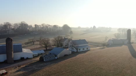 Mystic-farm-in-fog,-tranquil-countryside-aerial-scene-of-farmhouse-with-silo-and-sheds-in-morning-mist,-silhouettes-against-rising-sun