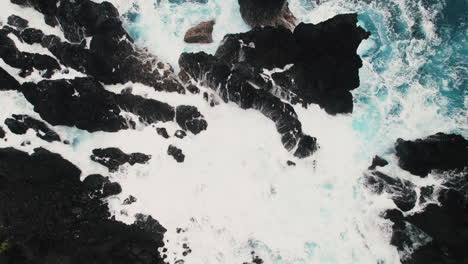 Aerial-top-down-view-of-blue-green-water-splashing-over-dark-lava-rocks-on-Hawaii-beach