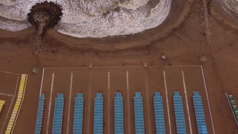 Aerial-top-down-of-colorful-tents-of-spa-resort-at-sandy-beside-crashing-waves-of-Ocean---Mar-del-Plata,Argentina---Ascending-drone-shot