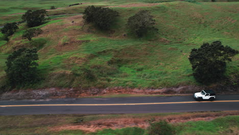 tracking-shot-of-white-jeep-driving-on-scenic-road-with-green-landscape---aerial-view