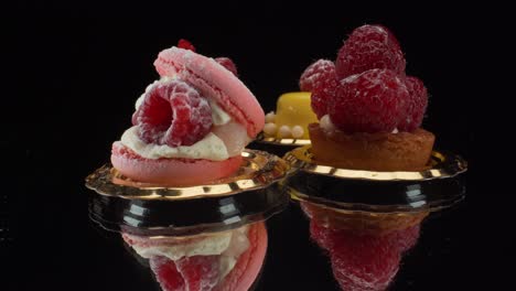 Macro-view-turning-colorful-pastries-on-black-reflective-glass-background,-luxury-patisserie,-mini-pavlova,-macaron,-raspberry-tart-and-mango-cake-4K-shot