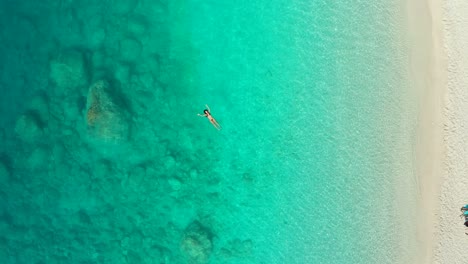 Aerial-ascending-view-of-young-woman-swimming-in-transparent-turquoise-sea-in-Greece,-summer-seascape-with-girl,-beach,-beautiful-blue-water,-top-view-from-drone
