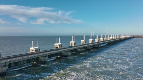 Aerial-slow-motion-shot-of-an-open-Eastern-Scheldt-storm-surge-barrier-and-wind-turbines-in-Zeeland,-the-Netherlands-on-a-beautiful-sunny-day-with-a-blue-sky