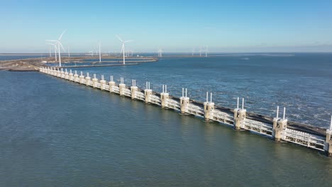 Aerial-slow-motion-shot-of-the-Eastern-Scheldt-storm-surge-barrier-and-wind-turbines-in-Zeeland,-the-Netherlands-on-a-beautiful-sunny-day