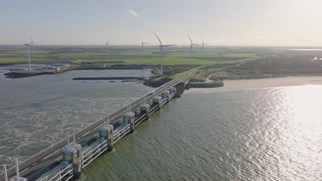 Aerial-slow-motion-shot-of-the-open-Eastern-Scheldt-storm-surge-barrier,-wind-turbines,-a-beach-and-farmland-on-a-beautiful-sunny-day-around-sunset