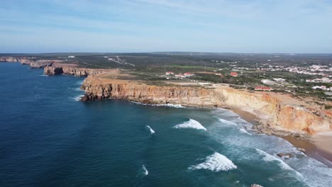 droneshot-of-the-beautiful-tonel-beach-with-big-waves-rolling-in-the-bay-and-sagres-on-the-hills,-southern-portugal-with-sunny-weather
