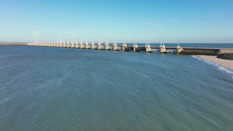 Aerial-slow-motion-shot-of-the-North-Sea,-the-Eastern-Scheldt-storm-surge-barrier-and-wind-turbines-in-Zeeland,-the-Netherlands-on-a-beautiful-sunny-day