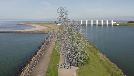 Aerial-shot-of-steel-artwork-on-a-dike-with-sluices-in-the-background-in-Lelystad,-the-Netherlands,-on-a-beautiful-sunny-summer-day