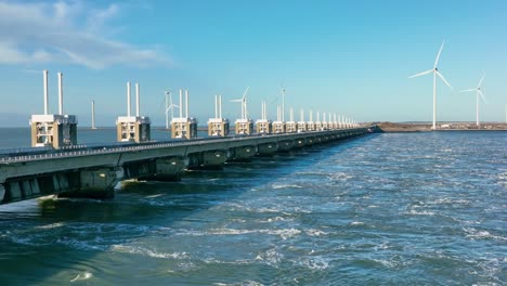 Aerial-slow-motion-shot-of-an-open-Eastern-Scheldt-storm-surge-barrier-and-wind-turbines-in-the-distance-in-Zeeland,-the-Netherlands-on-a-beautiful-sunny-day