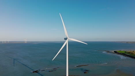 Aerial-slow-motion-shot-of-a-wind-turbine-along-the-coast-against-a-blue-sky-on-a-beautiful-sunny-day