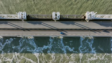 Aerial-overhead-shot-of-water-flowing-through-an-open-Eastern-Scheldt-storm-surge-barrier,-and-traffic-driving-over-it,-on-a-beautiful-sunny-day-in-Zeeland,-the-Netherlands