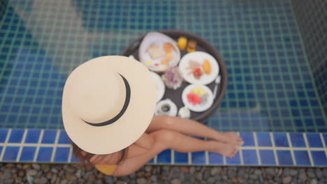 Top-view-on-the-gorgeous-slim-girl-in-straw-hat-sitting-poolside-with-her-legs-tucked-up-beneath-one,-lunch-set-up-next-to-the-pool