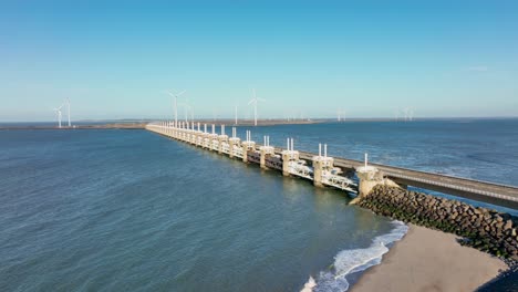 Aerial-slow-motion-shot-of-an-open-Eastern-Scheldt-storm-surge-barrier-and-wind-turbines-in-Zeeland,-the-Netherlands-on-a-beautiful-sunny-day-with-a-blue-sky