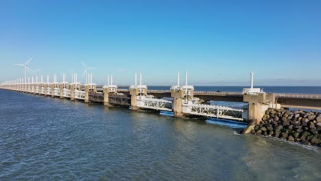 Aerial-slow-motion-shot-of-the-Eastern-Scheldt-storm-surge-barrier-and-wind-turbines-in-Zeeland,-the-Netherlands-on-a-beautiful-sunny-day-with-a-blue-sky