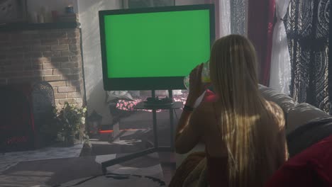 Green-screen-television-on-misty-hazy-morning-with-blonde-women-lady-girl-sitting-on-couch-cuddled-up-with-blanket-watching-and-drinking-from-wine-champagne-water-glass-and-checking-her-phone