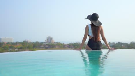 Slim-model-wearing-black-and-white-swimming-suit-and-black-hat-sitting-on-the-side-of-the-infinity-pool-with-her-back-to-the-camera,-city-view-on-the-background