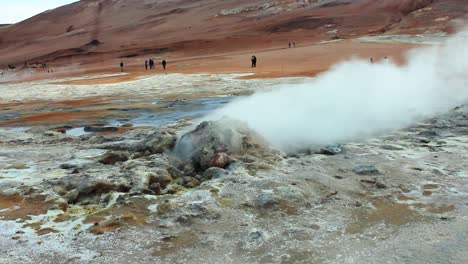A-smoking-volcanic-geyser,-with-people-and-sandy-brown-mountains-in-the-background,-Full-HD